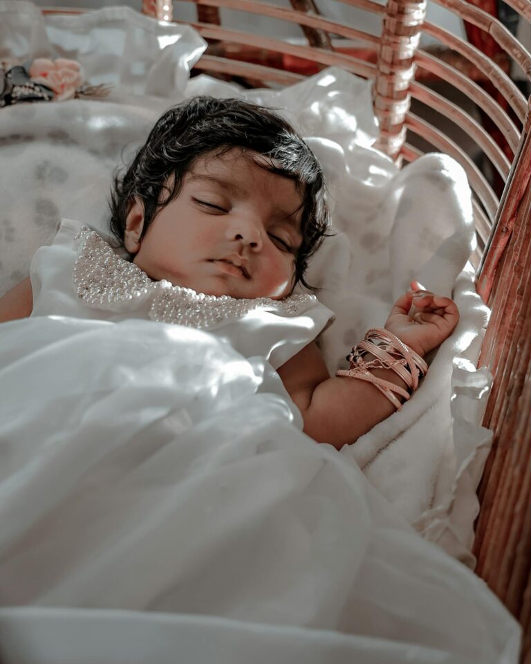 A serene scene of a baby sleeping in a wicker basket, wearing a white dress and bangles.